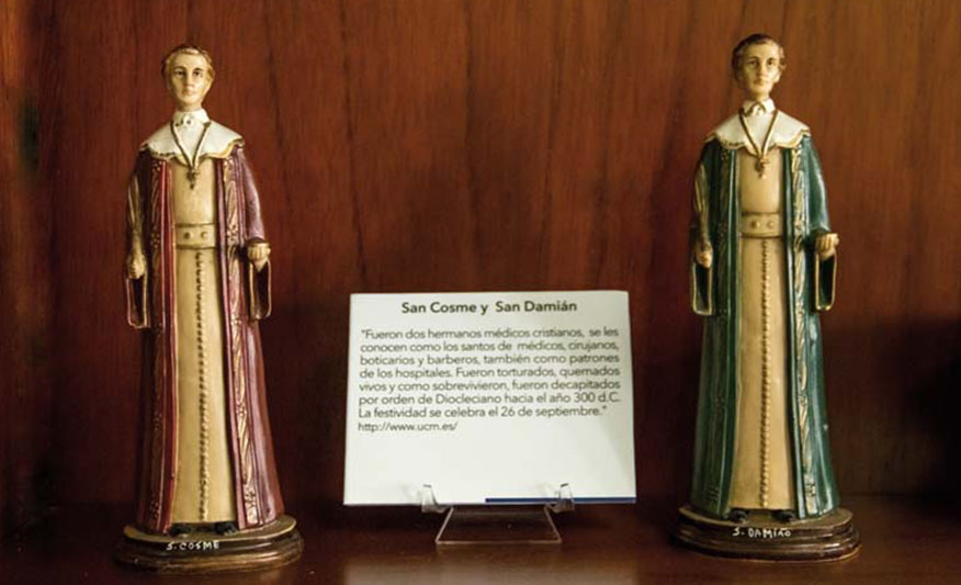 2 estatuillas de San Cosme y San Damián separadas por un texto colocado en medio de ellos que explica quiénes son. Los santos tienen la misma cara porque eran gemelos. Usan túnicas que les llegan hasta los pies. Una túnica es rosada y la otra verde. Cada uno de ellos lleva un crucifijo en el cuello